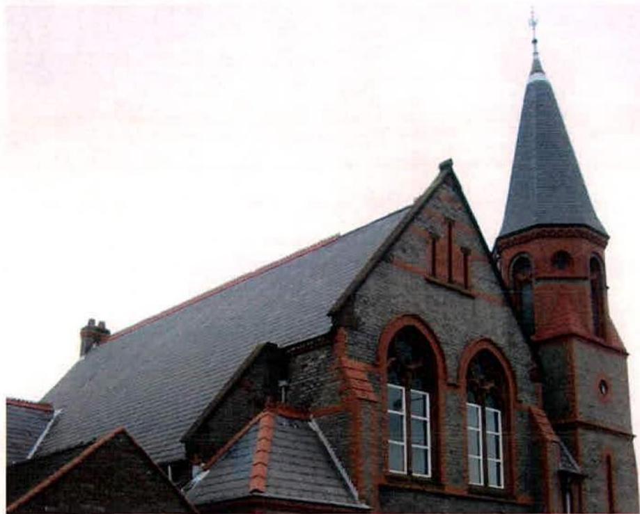 A photograph showing the upper exterior of a historic stone and red brick building featuring a slate roof, arched windows, and a prominent spire. The structure appears to be a registered building, likely a church or c...