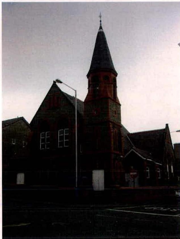 A low-light exterior photograph of a large stone building featuring a prominent tower with a spire, likely a church or chapel.