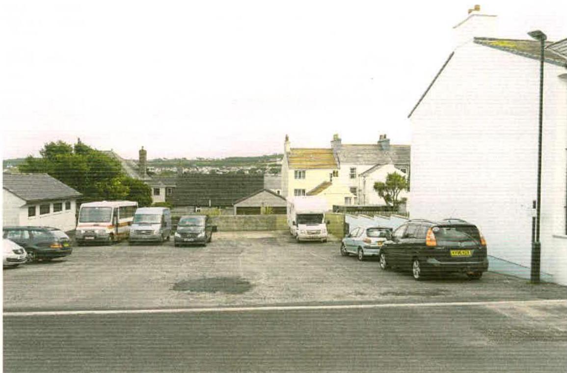 A photograph showing a paved parking area with several vans and cars, bordered by white buildings and a road in the foreground.