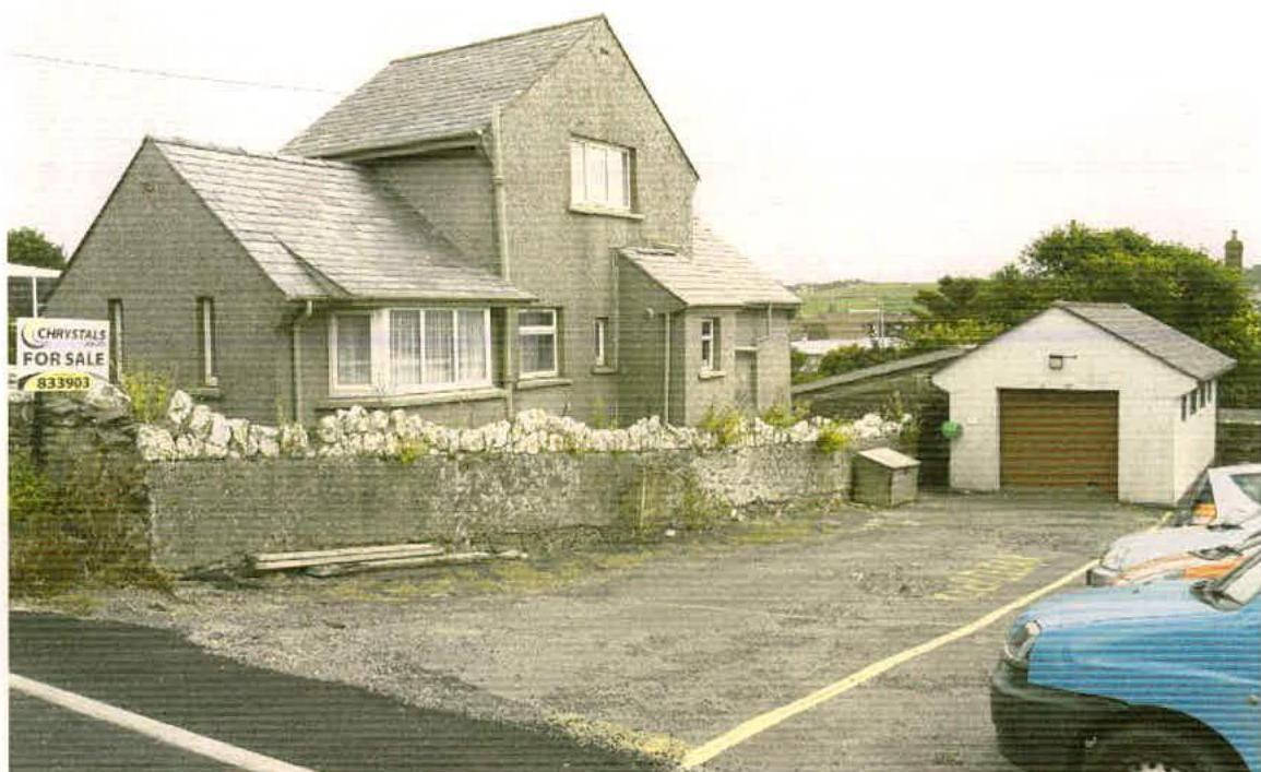 A photograph of a two-story detached stone house with a 'For Sale' sign, a detached garage, and a gravel parking area.