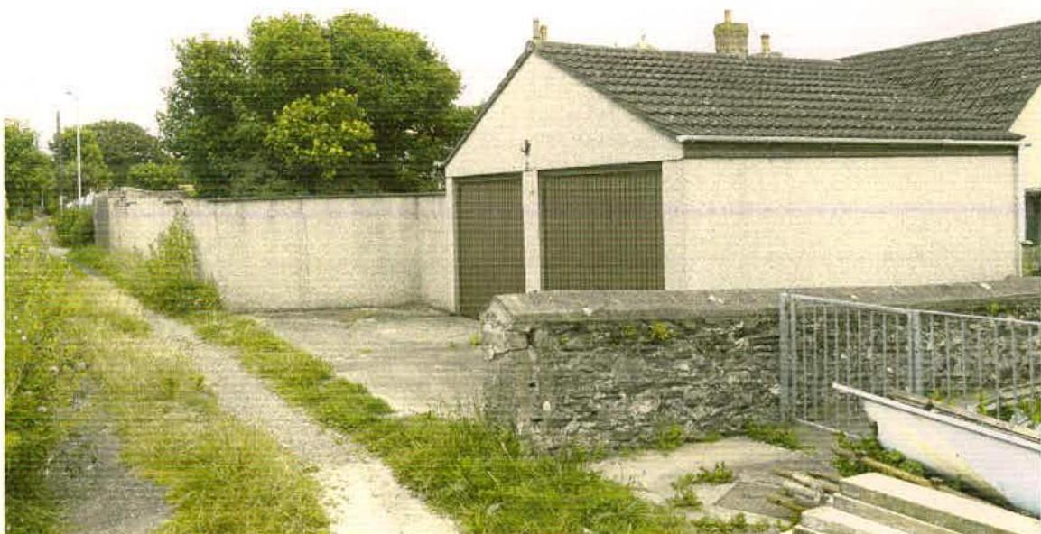 A photograph showing an existing single-story building with two large garage doors, situated next to a concrete wall and driveway.