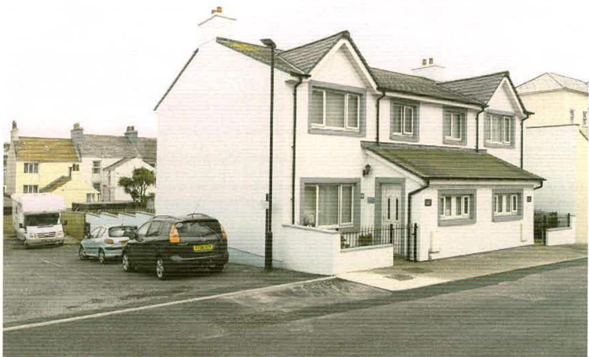 A photograph showing a white, two-story detached house with parked cars in front, representing the existing property subject to the application.