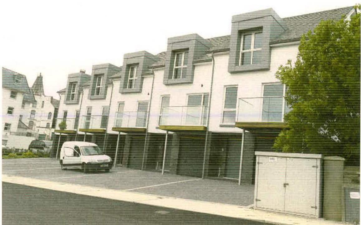 A photograph showing a row of six modern terraced houses with white facades, grey slate roofs, and balconies overlooking a parking area.