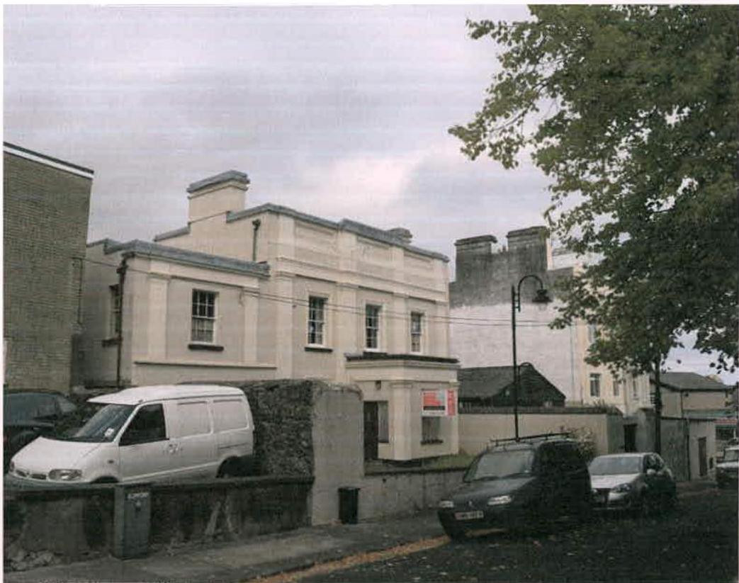 A street-level photograph showing a white two-story building with a stone wall and parked vehicles in the foreground.