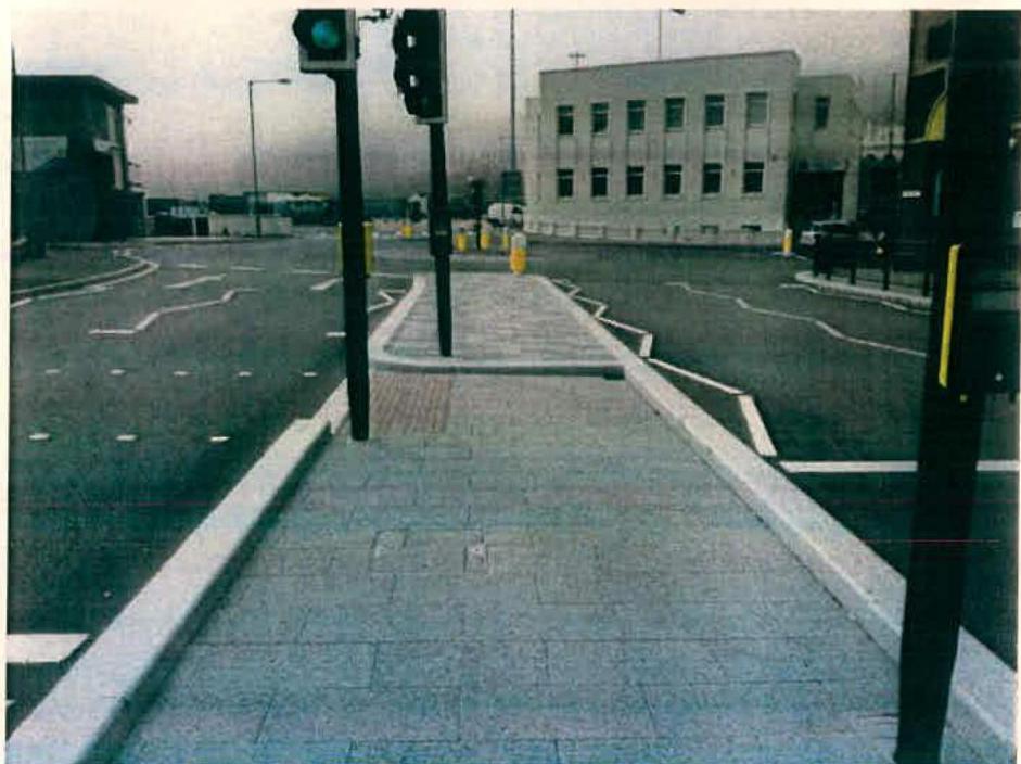 A photograph showing a paved pedestrian footway with traffic lights and a white commercial building in the background.