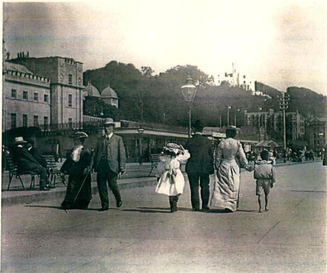 A vintage sepia-toned photograph showing pedestrians walking along a wide paved promenade or street with large buildings and a hill in the background.