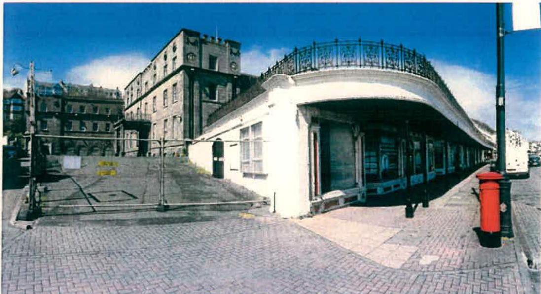 A street photograph showing a paved footway in the foreground, a white curved structure with a railing, and a large historic stone building in the background with a red post box nearby.
