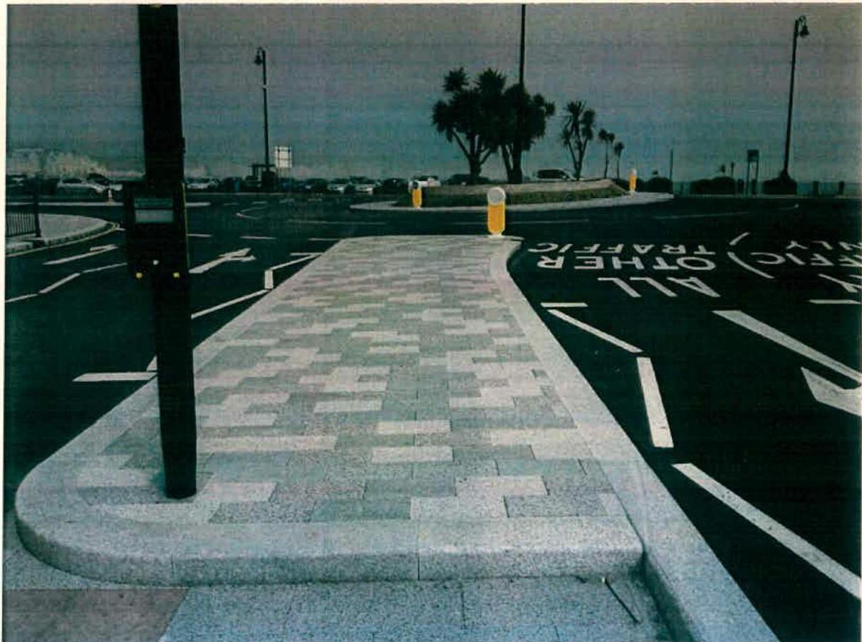 A photograph showing a newly paved footway with patterned stone slabs adjacent to a road with traffic markings and bollards.