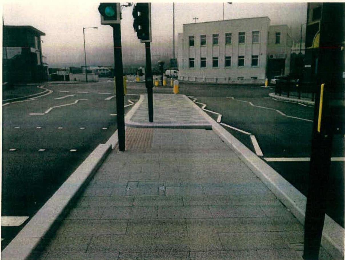 A street-level photograph showing a newly paved footway with traffic lights and a white building in the background.