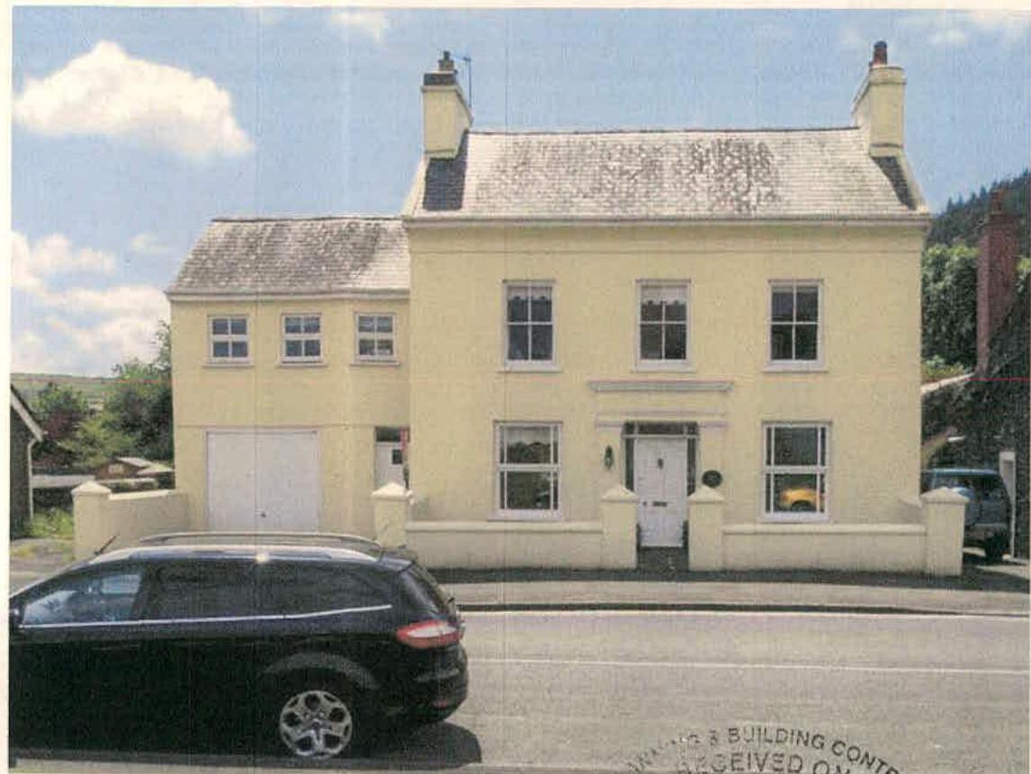 A photograph showing the exterior of a two-story cream-colored building with a slate roof and an attached garage wing. The building features sash windows and is situated on a street with a car parked in the foreground.