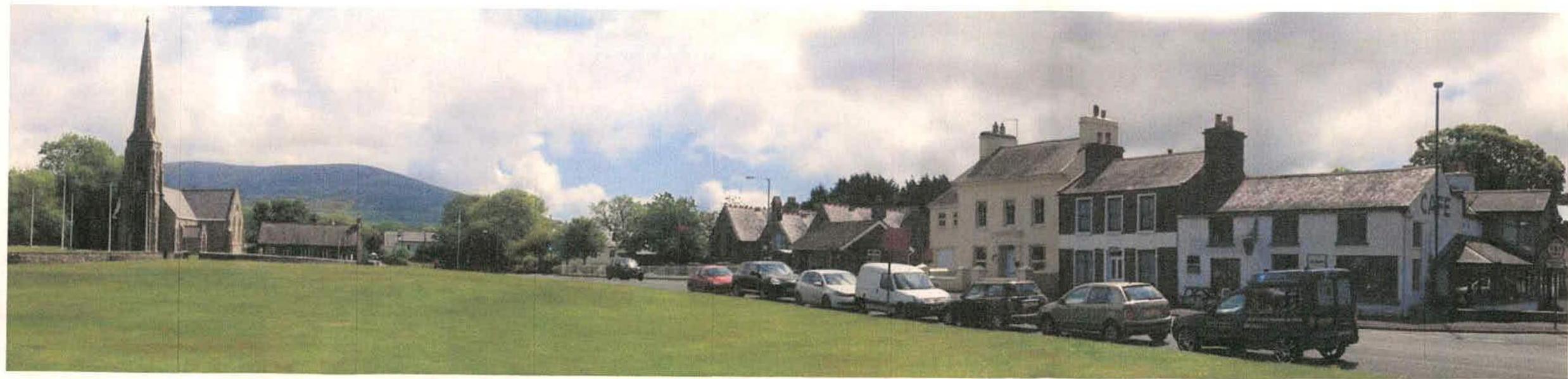 A wide panoramic photograph showing a village scene with a church spire on the left, a grassy green, and a row of stone buildings with parked cars on the right.