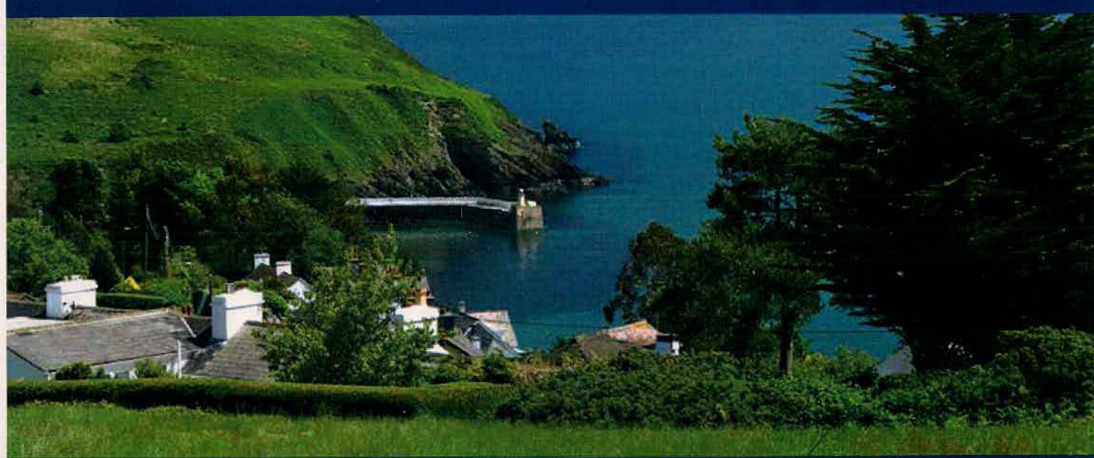 A scenic photograph showing a coastal landscape with a small harbor, green hills, and rooftops of buildings in the foreground.