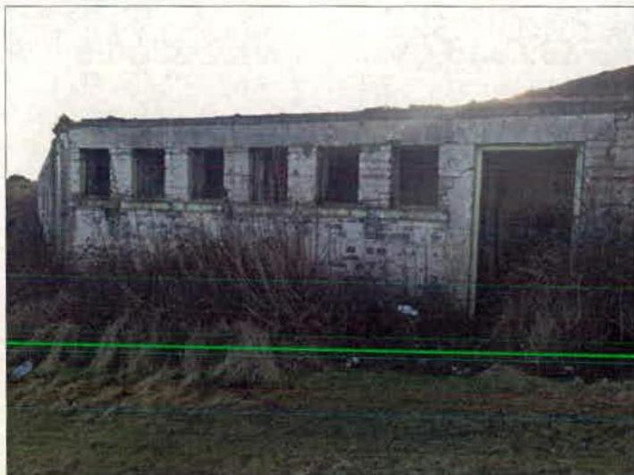 A photograph showing a dilapidated, single-story concrete building with empty window openings and overgrown vegetation.
