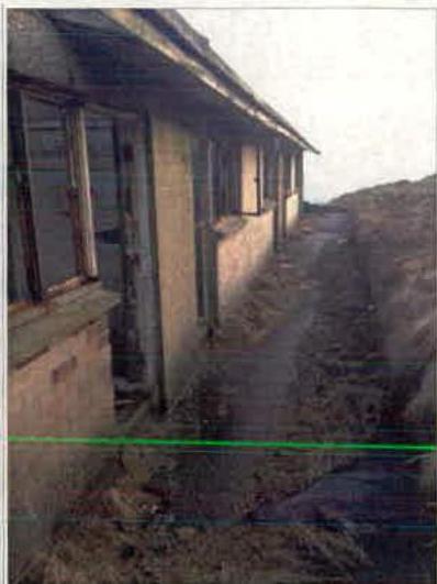 A photograph showing the side elevation of a long, single-story building with multiple windows, situated on rough, uneven ground.