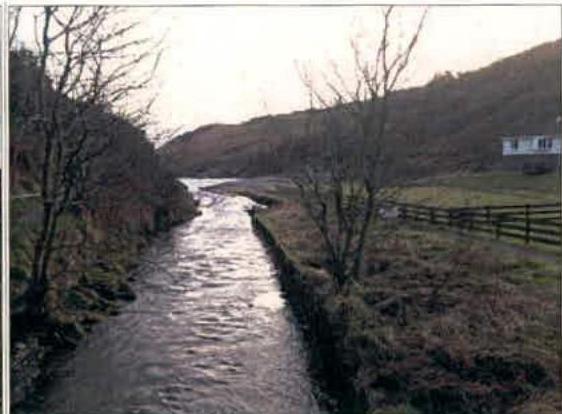 A photograph showing a stream flowing through a rural landscape with a white house visible on the hillside in the background.
