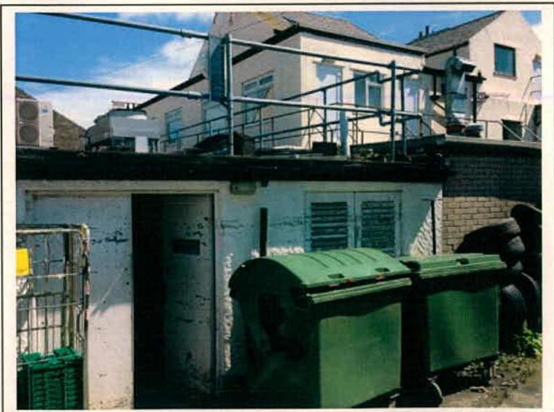 A photograph showing the exterior of a white building with a flat roof, featuring a doorway and green waste bins in the foreground.