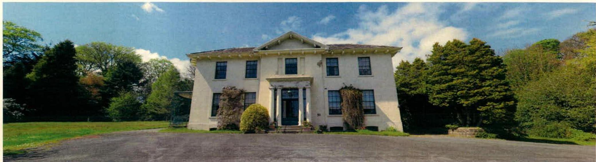 A wide photograph showing a large, white, two-story detached house with a classical entrance facade, set within a green, tree-lined site with a driveway in the foreground.