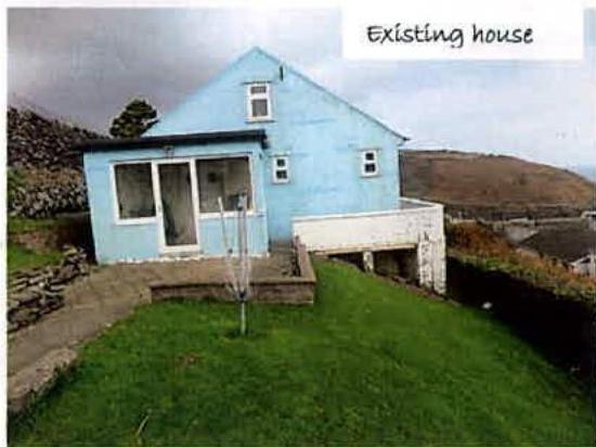 A photograph of the existing light blue single-story house with a white extension, situated on a grassy slope with hills in the background.