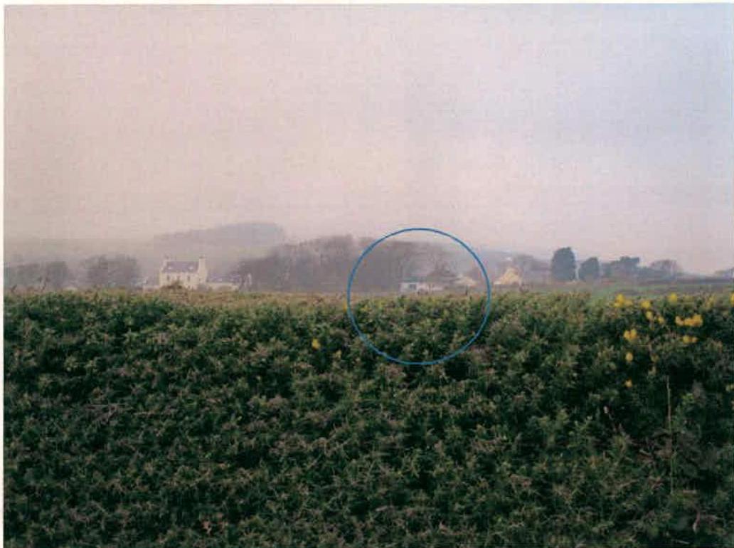 A photograph showing a rural landscape with a field of crops in the foreground and a blue circle highlighting a specific site location in the distance near trees and buildings.