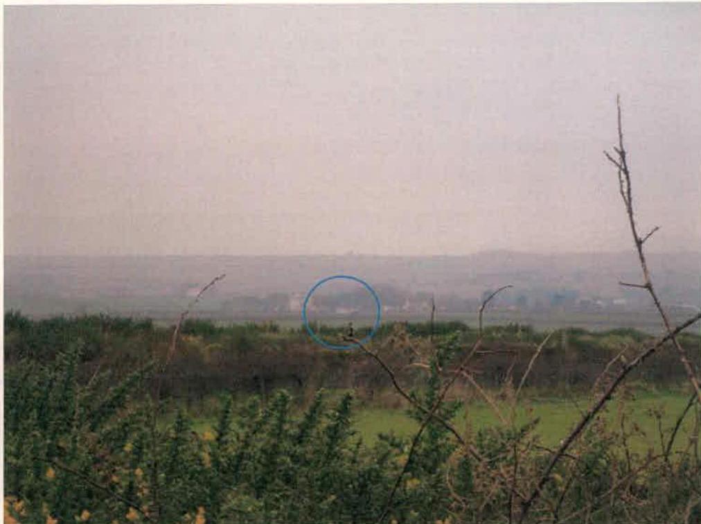 A distant landscape photograph showing a rural view with a blue circle highlighting a specific location in the distance, likely indicating the site context.