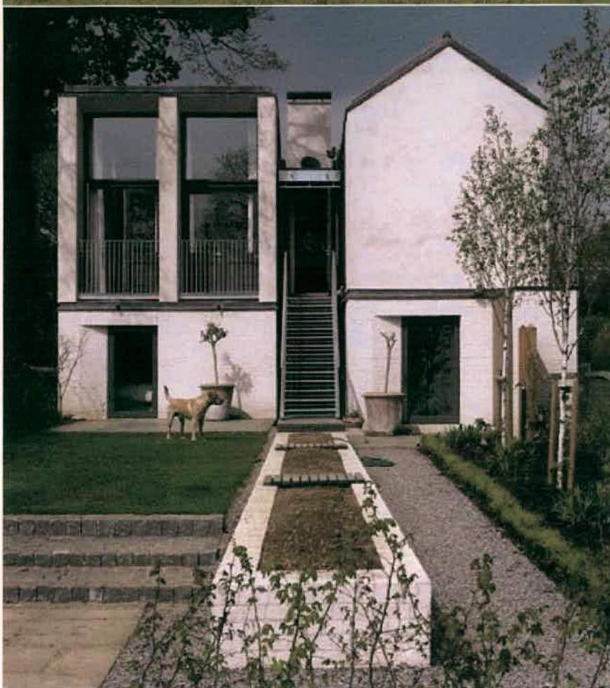 A photograph showing a modern two-story replacement dwelling with a mix of concrete and white rendered walls, featuring an external staircase and landscaped front garden.