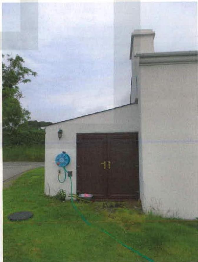 A photograph showing the exterior of a white rendered building with a large brown double door, likely a garage, situated next to a road in a rural setting.