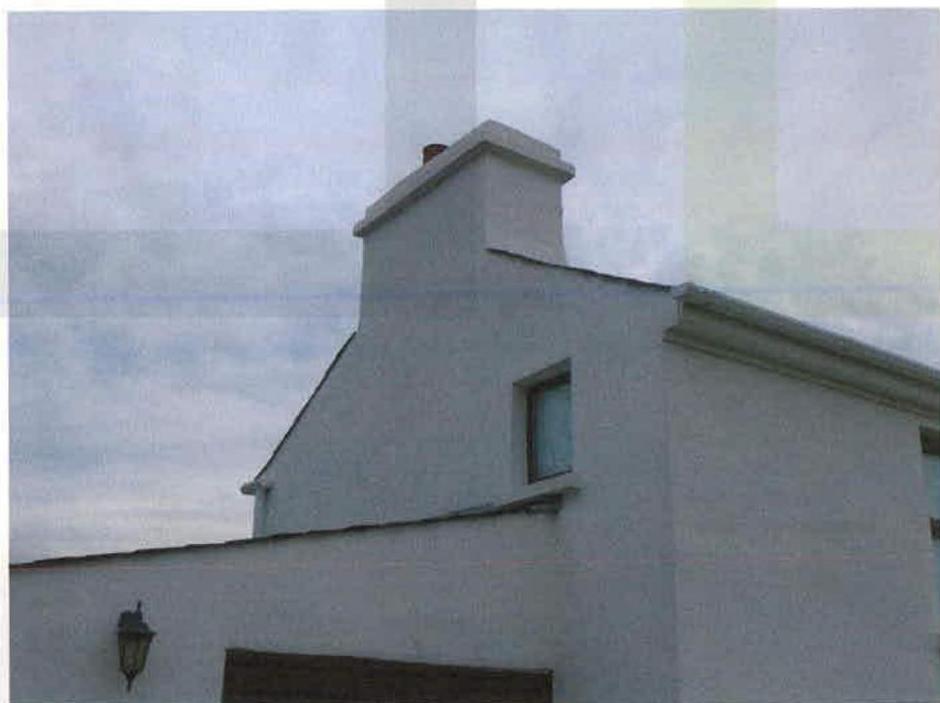 A low-resolution photograph showing the upper gable end and chimney stack of a white rendered building against a cloudy sky.