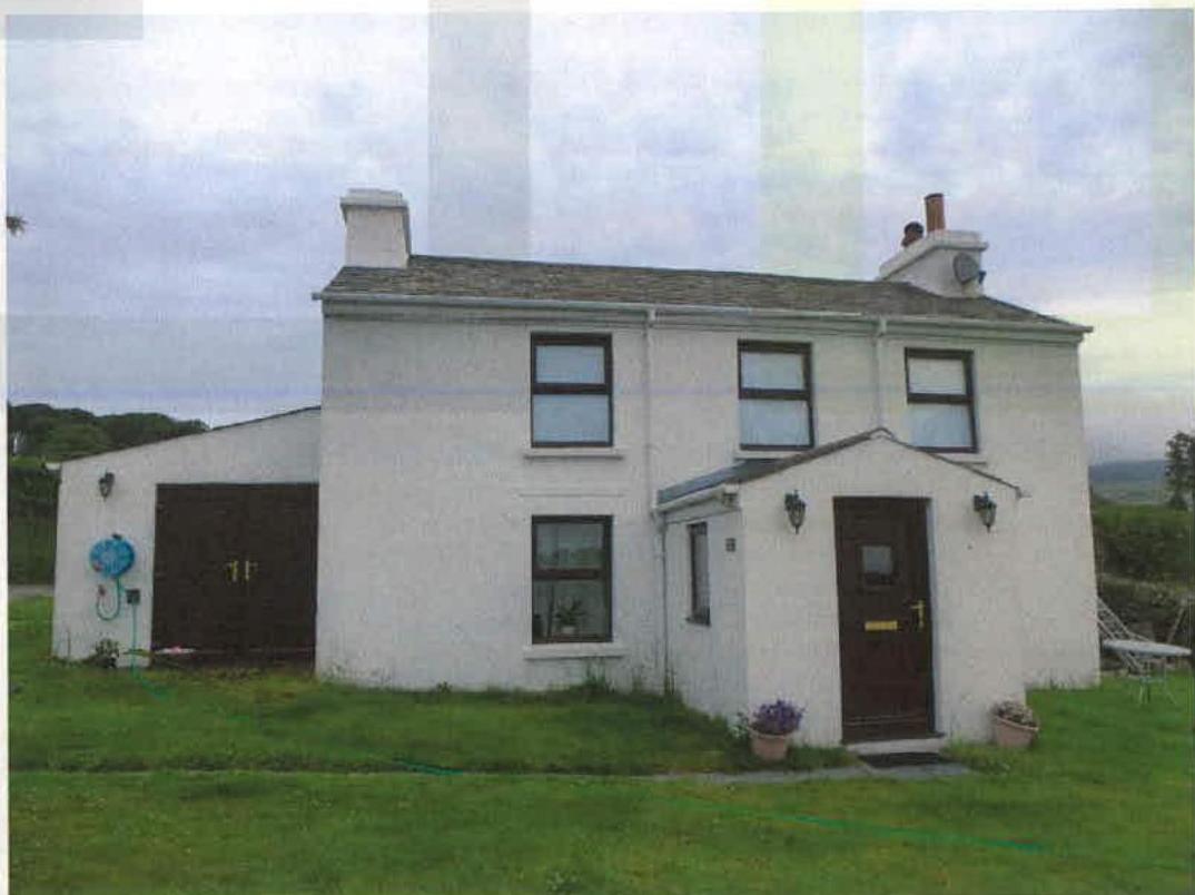 A photograph of a white, two-story detached house with a single-story garage extension on the left and a small porch on the right, situated in a rural grassy setting.