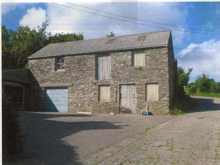 A photograph of a detached stone building, likely a barn, featuring boarded windows and a garage door with a paved driveway in front.