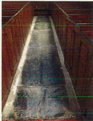 A grainy photograph showing a long, narrow concrete path or ramp flanked by wooden walls, likely depicting existing access conditions within a building.
