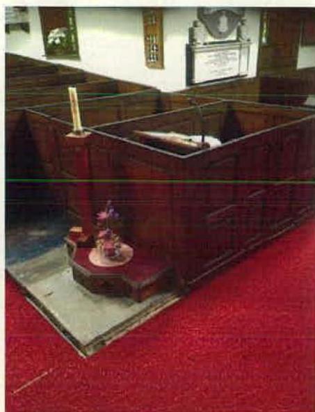 Interior photograph of a church showing wooden pews, a raised pulpit structure, and red carpeting.