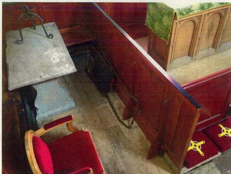 The image shows the interior of a historic building, likely a church or chapel, featuring wooden pews, a stone slab, and a red chair. It appears to be a condition survey photograph documenting the existing state befor...