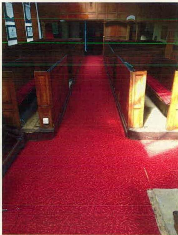 Interior photograph of a building, likely a church or chapel, showing wooden pews and a red carpeted aisle with green lines indicating a path.