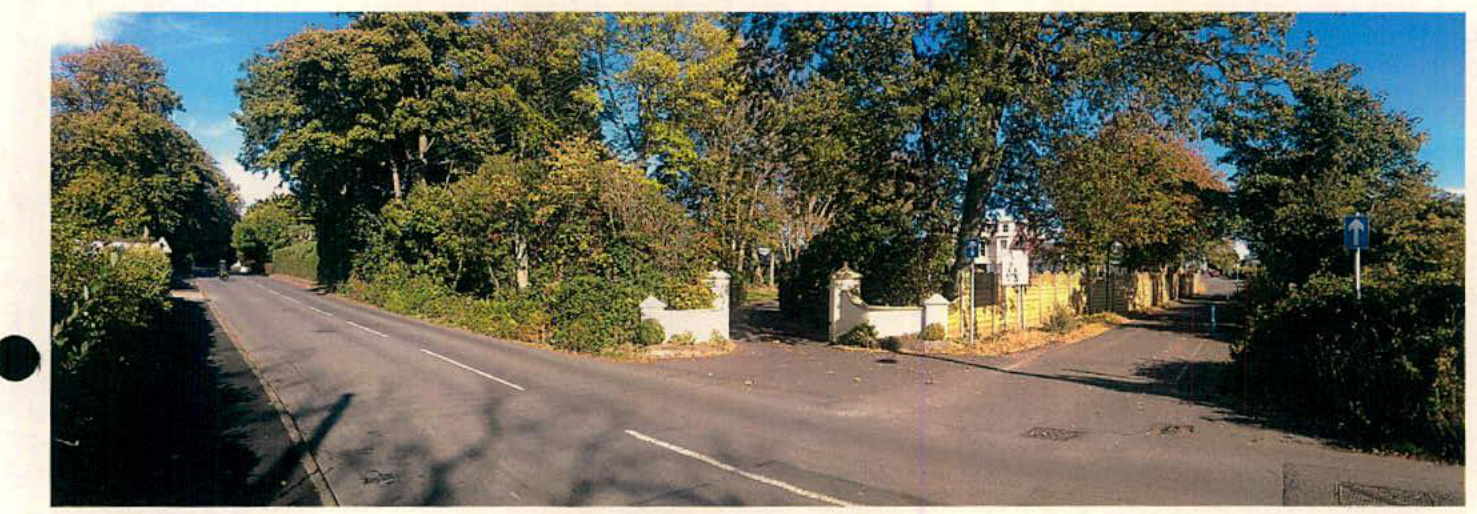A panoramic photograph showing a road junction with a driveway entrance on the right, flanked by trees and hedges, with a white house visible in the background.