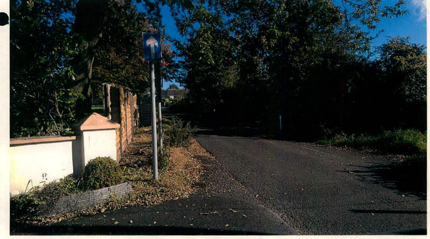 A photograph showing a paved access road or driveway entrance featuring a blue directional sign and a white boundary wall with brick pillars on the left.