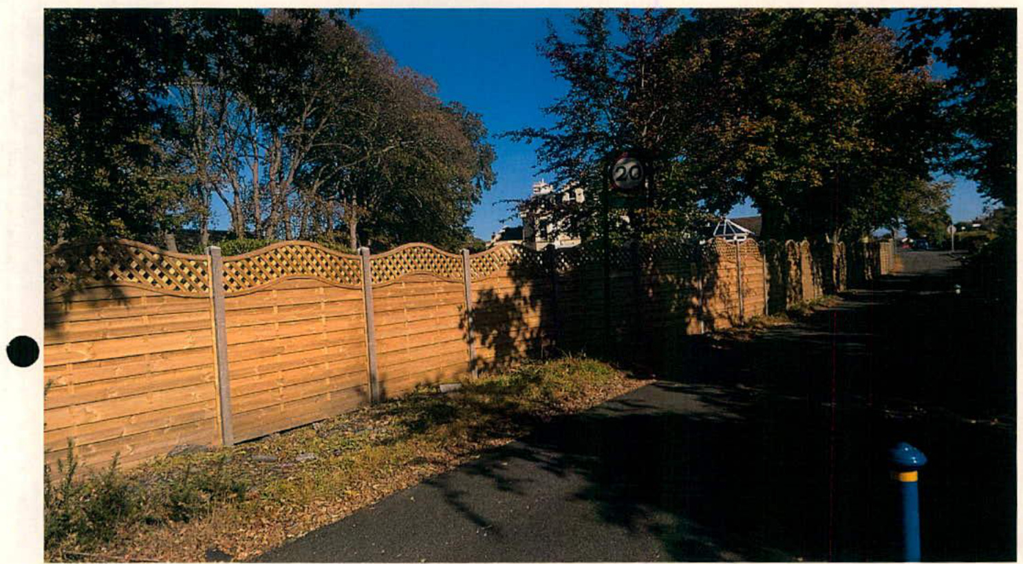 A photograph showing a wooden boundary fence with lattice detailing running alongside a paved path, with trees and a speed limit sign visible in the background.