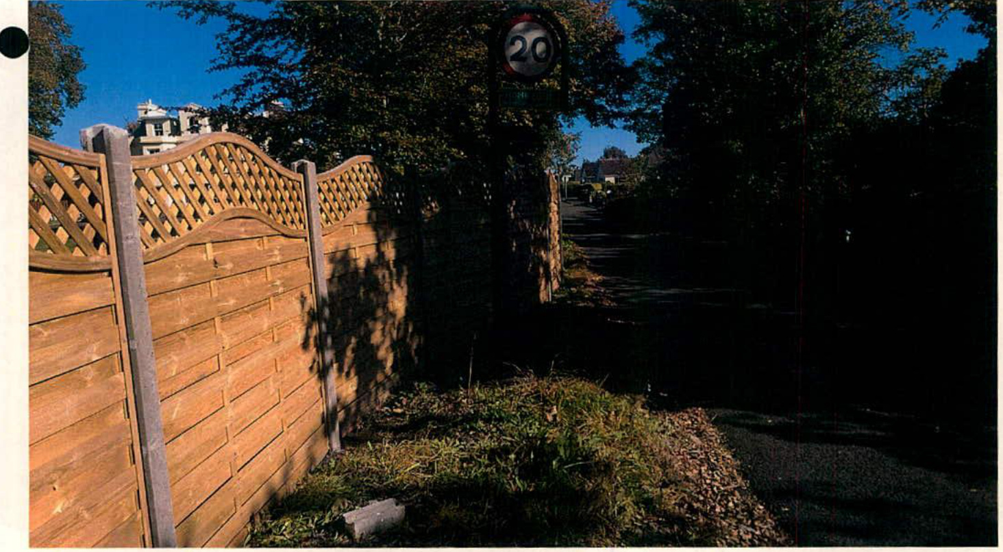 A photograph showing a wooden fence with lattice top running along a roadside, with a speed limit sign and trees in the background.