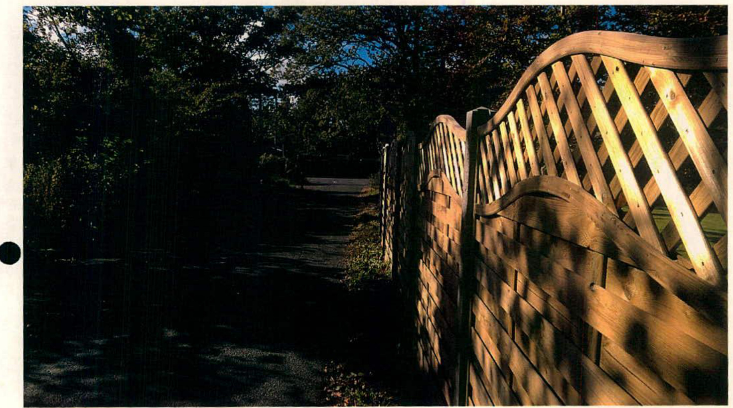 A photograph showing a wooden fence with a wavy top rail and lattice detailing running alongside a paved path or driveway leading into a wooded area.