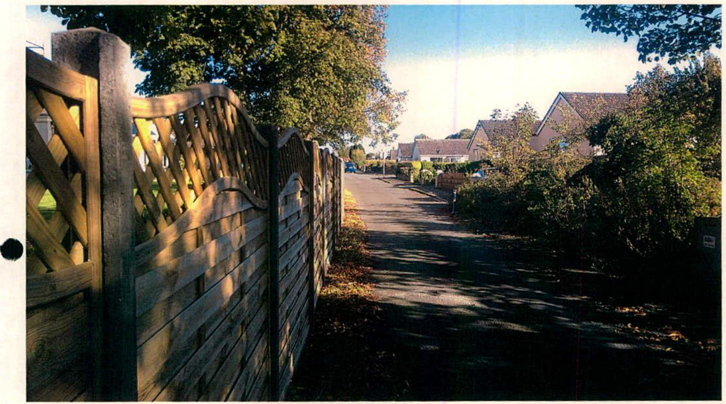 A photograph showing a wooden fence with lattice detailing along the left side of a residential road, with houses visible in the background.