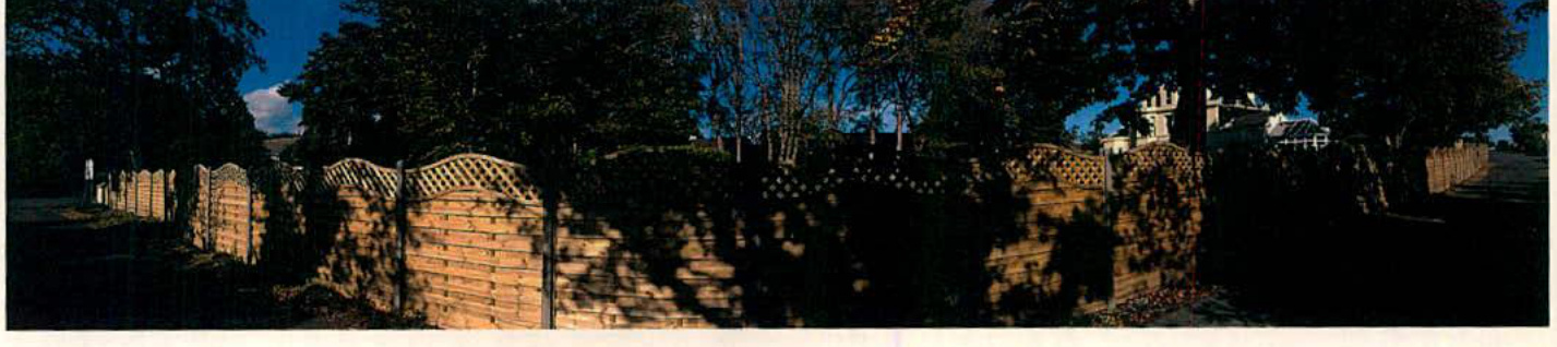A panoramic photograph showing a long wooden panel fence with decorative lattice tops running along a property boundary. Trees and foliage cast shadows across the fence, with a white house partially visible in the bac...