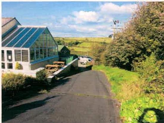 A photograph showing a white building with a large glass conservatory-style extension on the left, situated next to a driveway with parked vehicles in a rural setting.