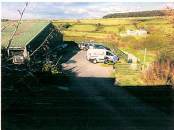 A photograph of a rural site featuring a large building with a green roof on the left and a driveway with parked vehicles.