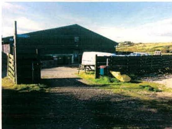 A photograph showing a large, dark agricultural building, likely a barn, situated in a rural setting with a wooden gate and fence in the foreground.