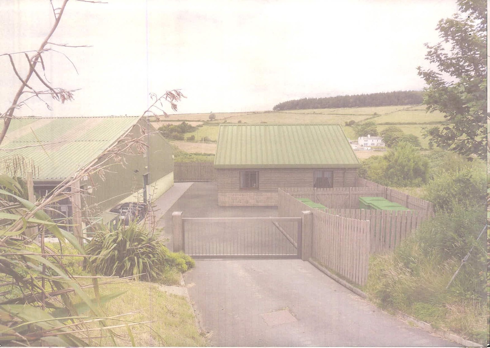 A photograph showing a rural property featuring a large agricultural barn and a smaller single-storey building with a green roof. A paved driveway leads to a metal gate and fenced enclosures in the foreground.