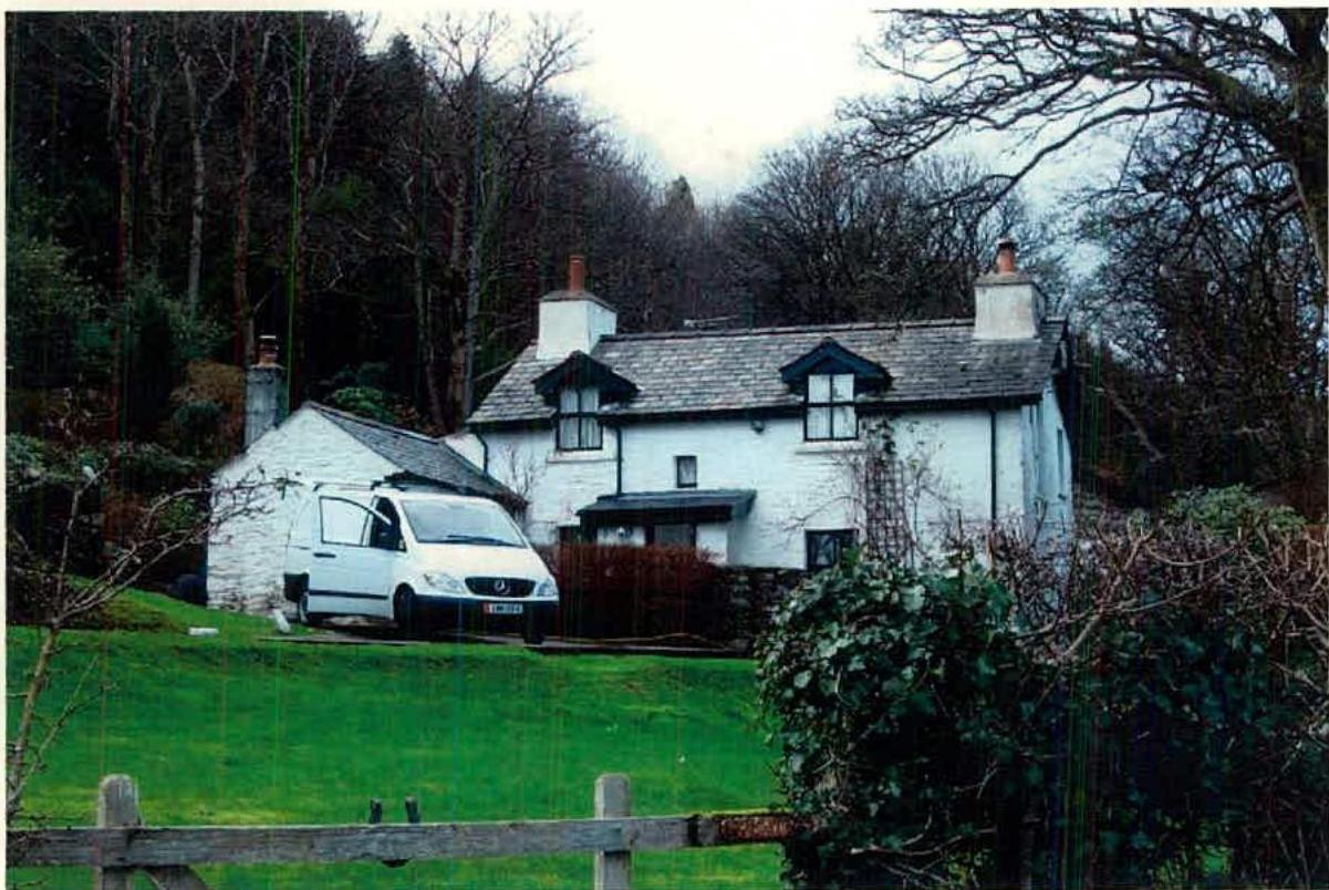 A photograph showing a white, detached house with a slate roof situated in a wooded, rural setting. A white van is parked on the driveway next to a smaller outbuilding on the left side of the property.