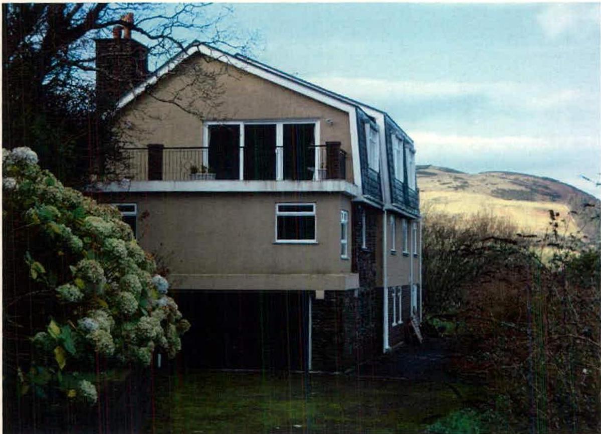 A photograph showing a two-story detached house with a balcony and dormer windows, situated in a rural setting with hills in the background.
