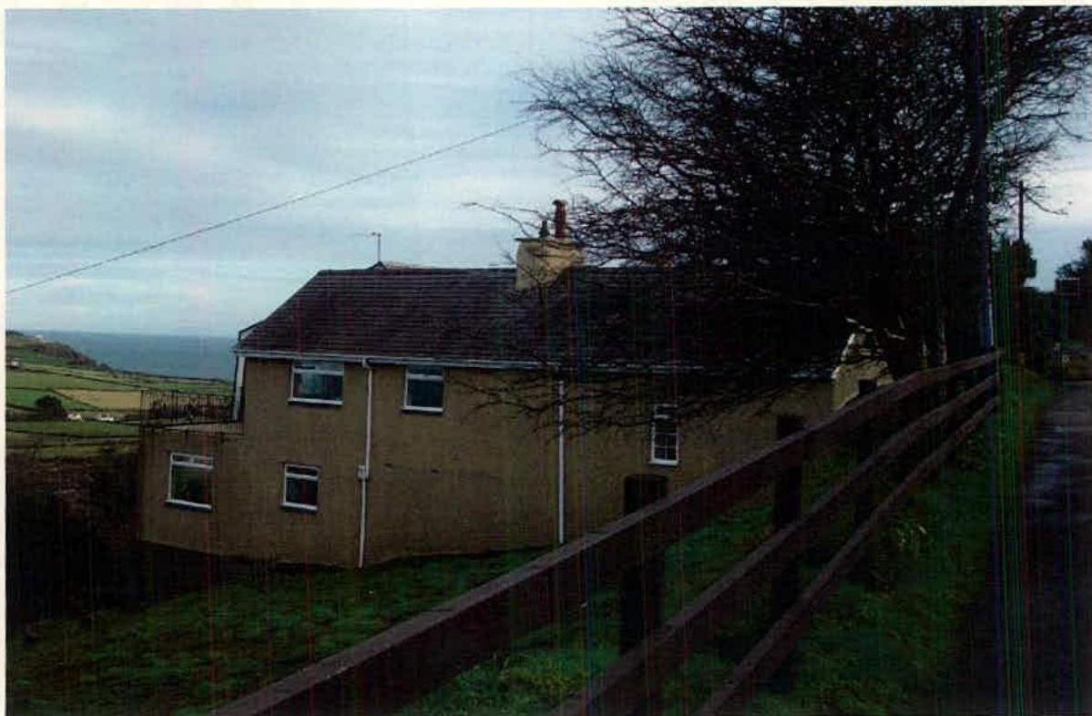 A photograph showing a two-story detached house situated on a slope with a view of the sea and fields in the background.