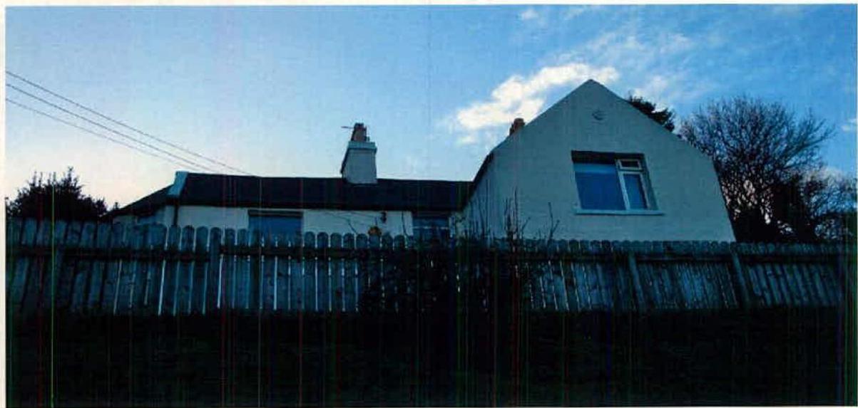 A photograph showing a white single-story detached house with a dark roof, viewed from behind a wooden fence in a rural setting.