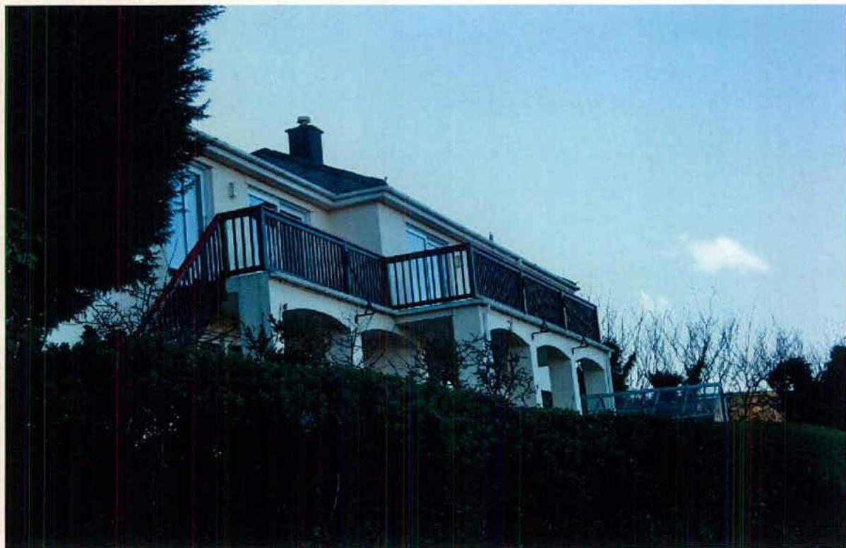 A low-angle photograph showing a two-story detached house with a prominent balcony and dark railing, situated on a slope with vegetation in the foreground.