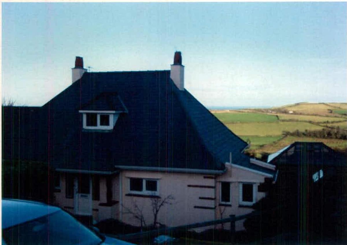 A photograph of a detached two-storey house with a dark slate roof and white chimneys, set against a backdrop of rural fields and the sea.